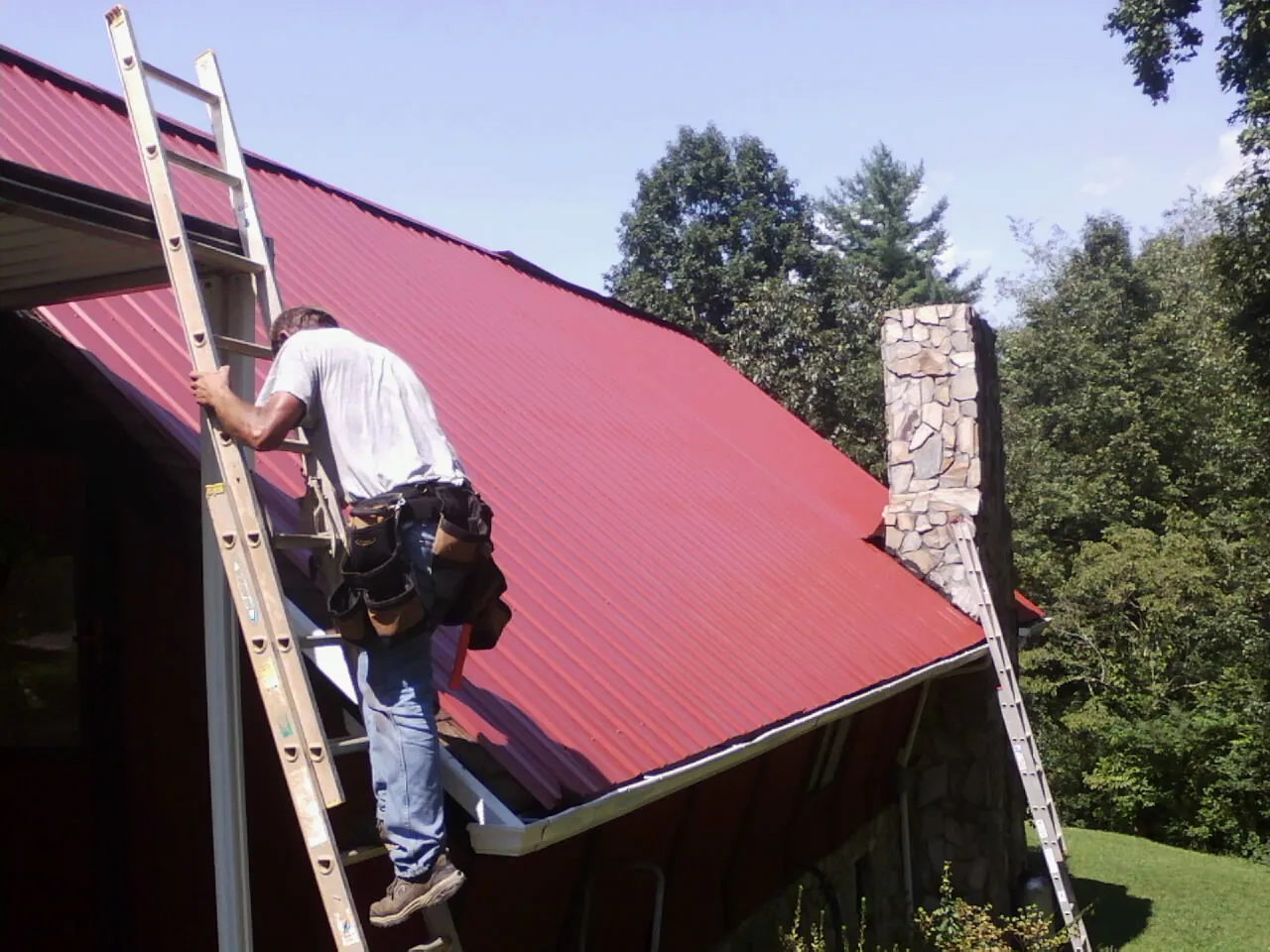 Roofer on a ladder beside a red metal roof and stone chimney — Soffit & Fascia Repair by Roofing Brothers in Wellsburg